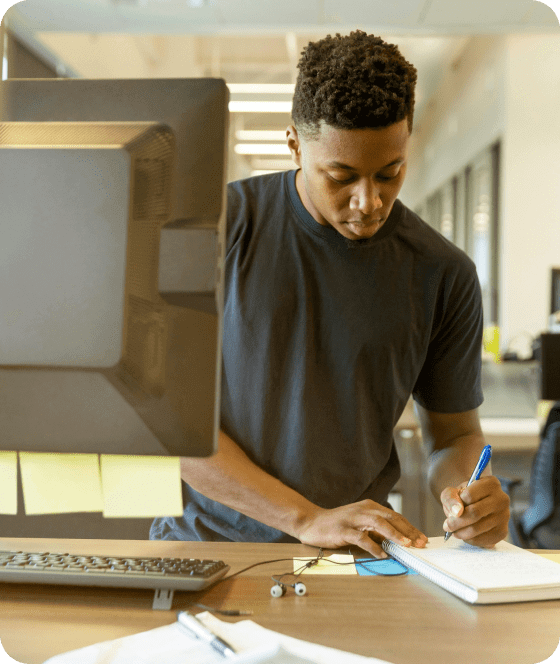 Homem em pé, de frente para um computador, fazendo anotações em um caderno